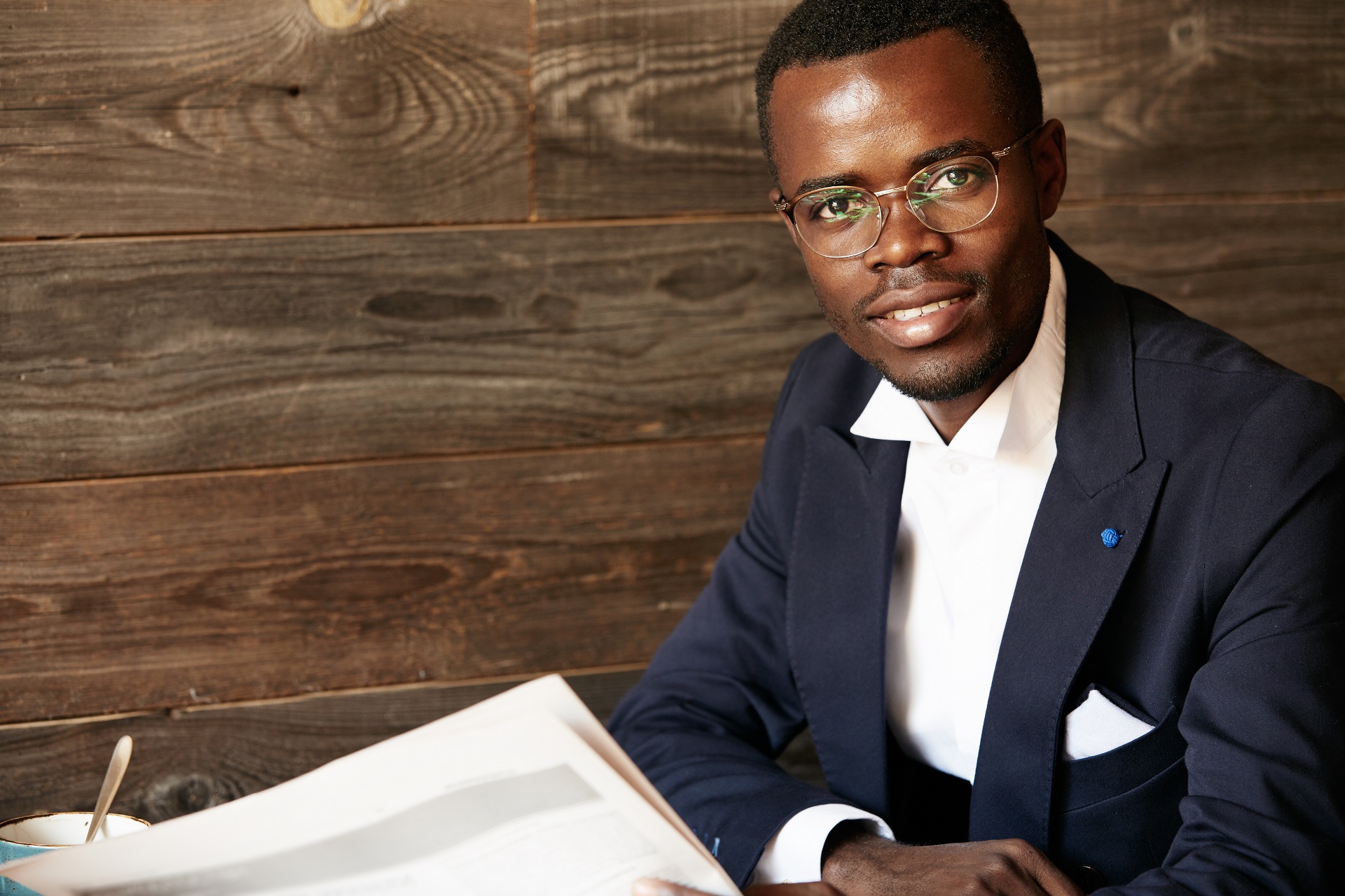 Lifestyle and people concept. Headshot of young African businessman in formal suit and glasses looking and smiling at the camera with confident and happy expression against wooden wall background